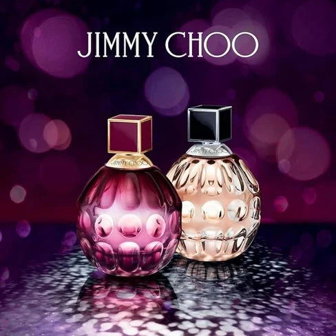 Two faceted perfume bottles, one pink with a gold cap and one pale with a black cap, are displayed on a reflective surface with the “JIMMY CHOO” logo above them on a purple bokeh background.