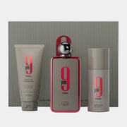 Afnan 9PM Rebel 100ml EDP Gift Set by Afnan, including shower gel, unisex Eau De Parfum spray, and deodorant—each in coordinated gray and red packaging—presented in front of a chic striped box.