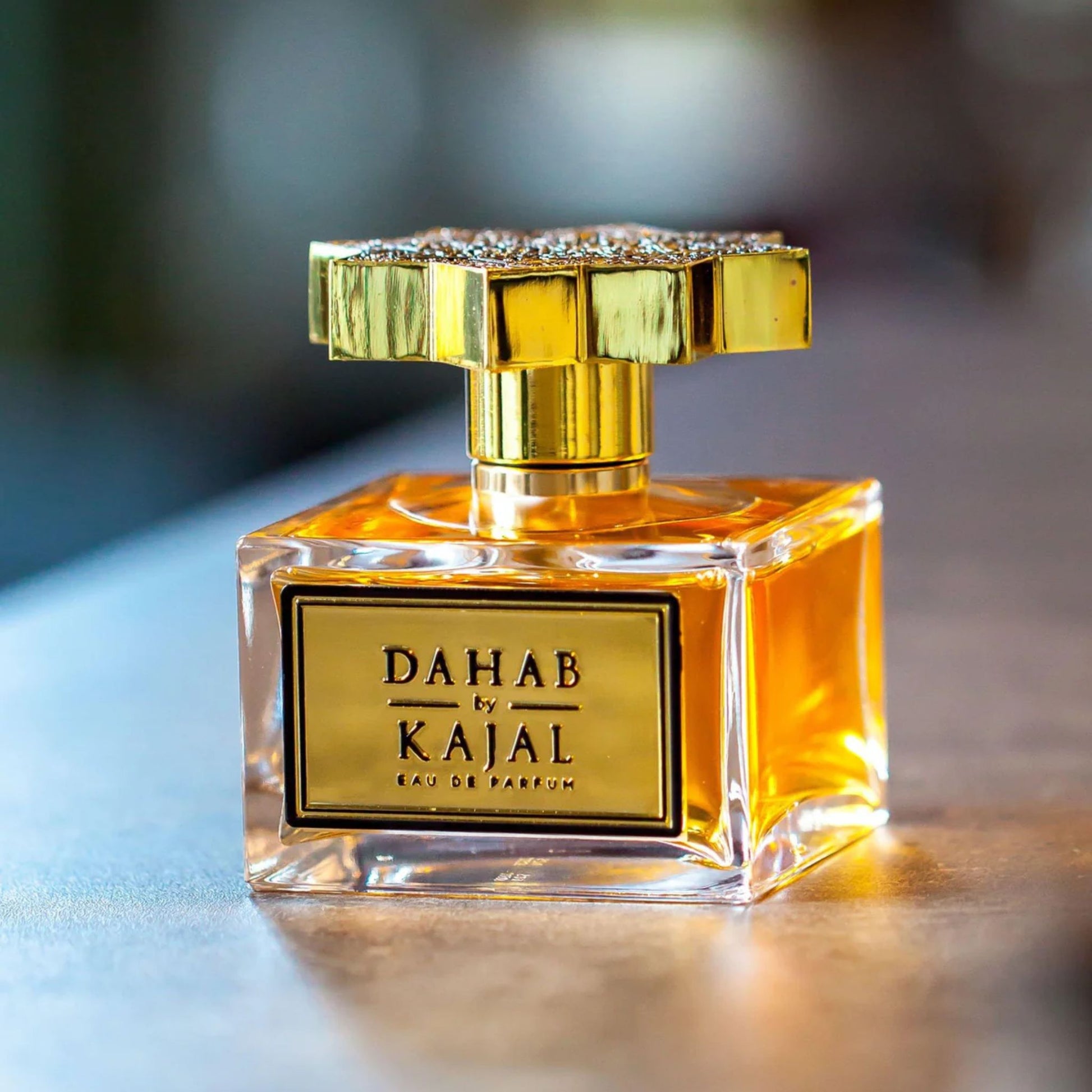 A square glass bottle of Dahab Kajal by Kajal, a mesmerizing women’s fragrance, featuring a gold label and ornate gold cap, rests on a smooth surface.