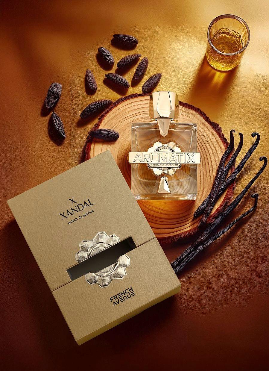 A bottle of French Avenue XXandal Aromatix 100ml Extrait De Parfum by French Avenue beside its open box, with vanilla beans, tonka beans, and a glass of amber liquid on wood—a welcoming unisex floral fragrance.