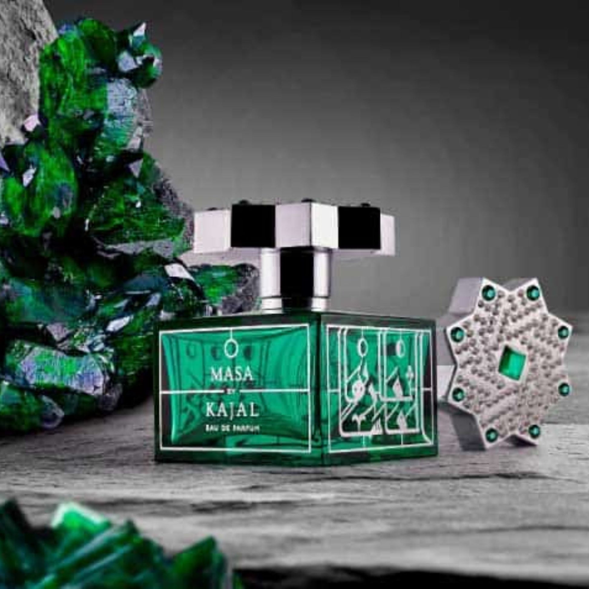 A green bottle of Masa Kajal by Kajal is displayed with its jeweled cap off, surrounded by green crystals and stones in a studio setting, highlighting this elegant unisex fragrance.
