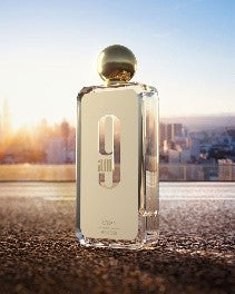 Afnan 9AM White 100ml EDP by Afnan, in a clear glass bottle with a gold cap, sits on a surface before a cityscape at sunrise—a refreshing men’s fragrance.