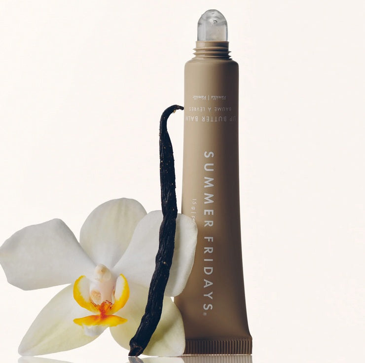 A tube of Summer Fridays Lip Butter Balm Vanilla stands upright next to a white orchid flower and a vanilla bean on a light background.
