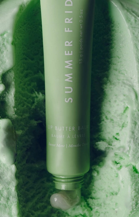A green tube of Summer Friday Lip Butter Balm Sweet Mint rests on a mint-textured surface, with a small amount of this vegan balm dispensed from the tip.