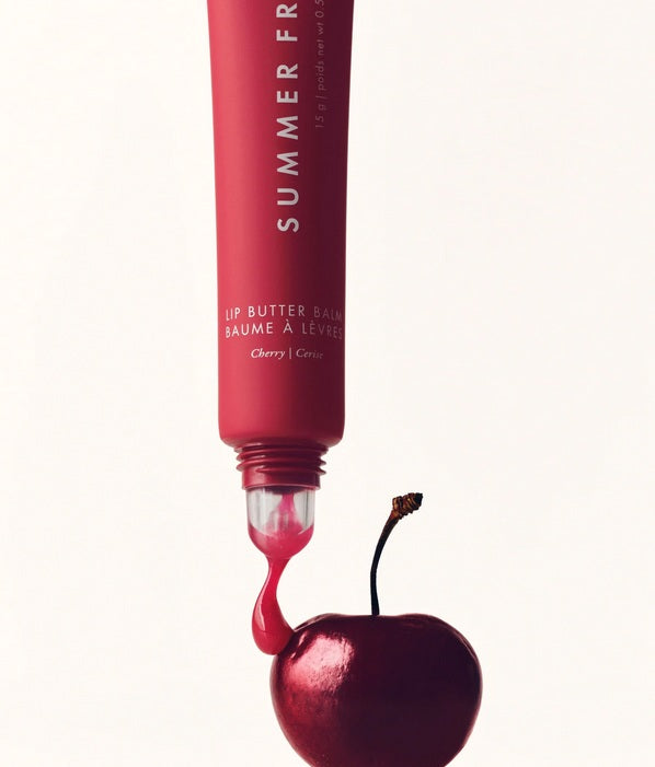 A tube of Summer Fridays Lip Butter Balm Cherry dispenses product onto the stem of a real cherry, with a plain background.