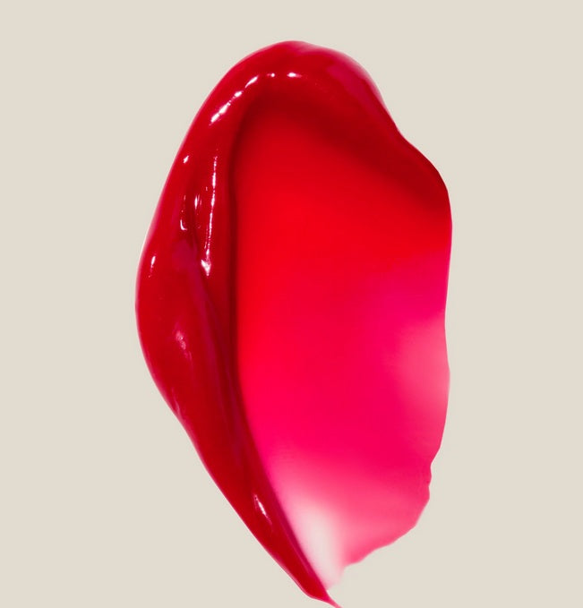 A close-up of a smear of Summer Fridays Lip Butter Balm Cherry, a glossy bright pink and red nourishing lip mask, on a plain beige background.