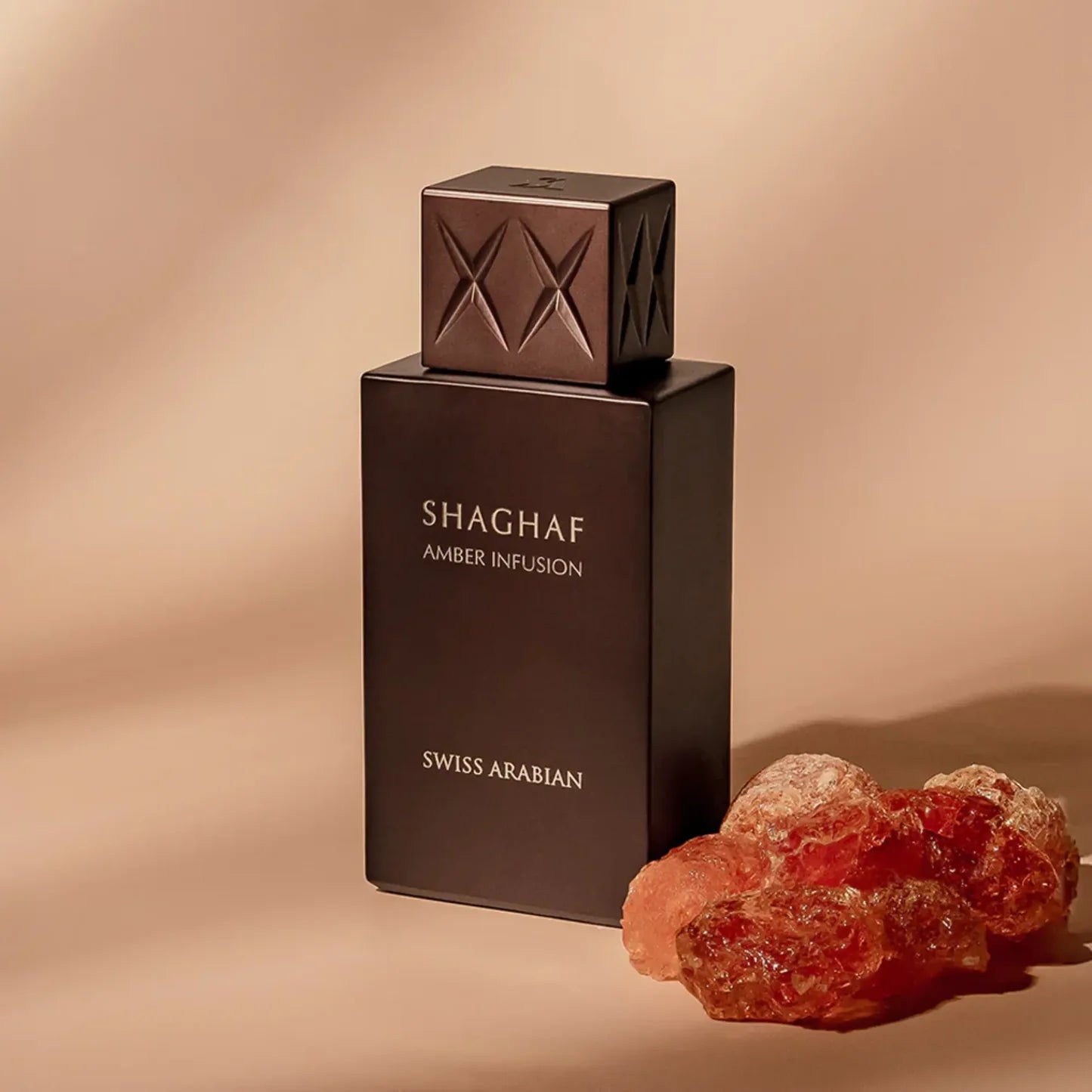 A dark brown rectangular bottle of Swiss Arabian Shaghaf Amber Infusion 75ml EDP stands beside amber resin on a beige surface, showcasing the alluring amber fragrance from Swiss Arabian.