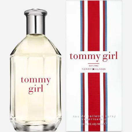 A clear 100ml Tommy Girl EDT bottle by Tommy Hilfiger stands next to a red, white, and blue striped box, reflecting a vibrant floral fruity fragrance for women.