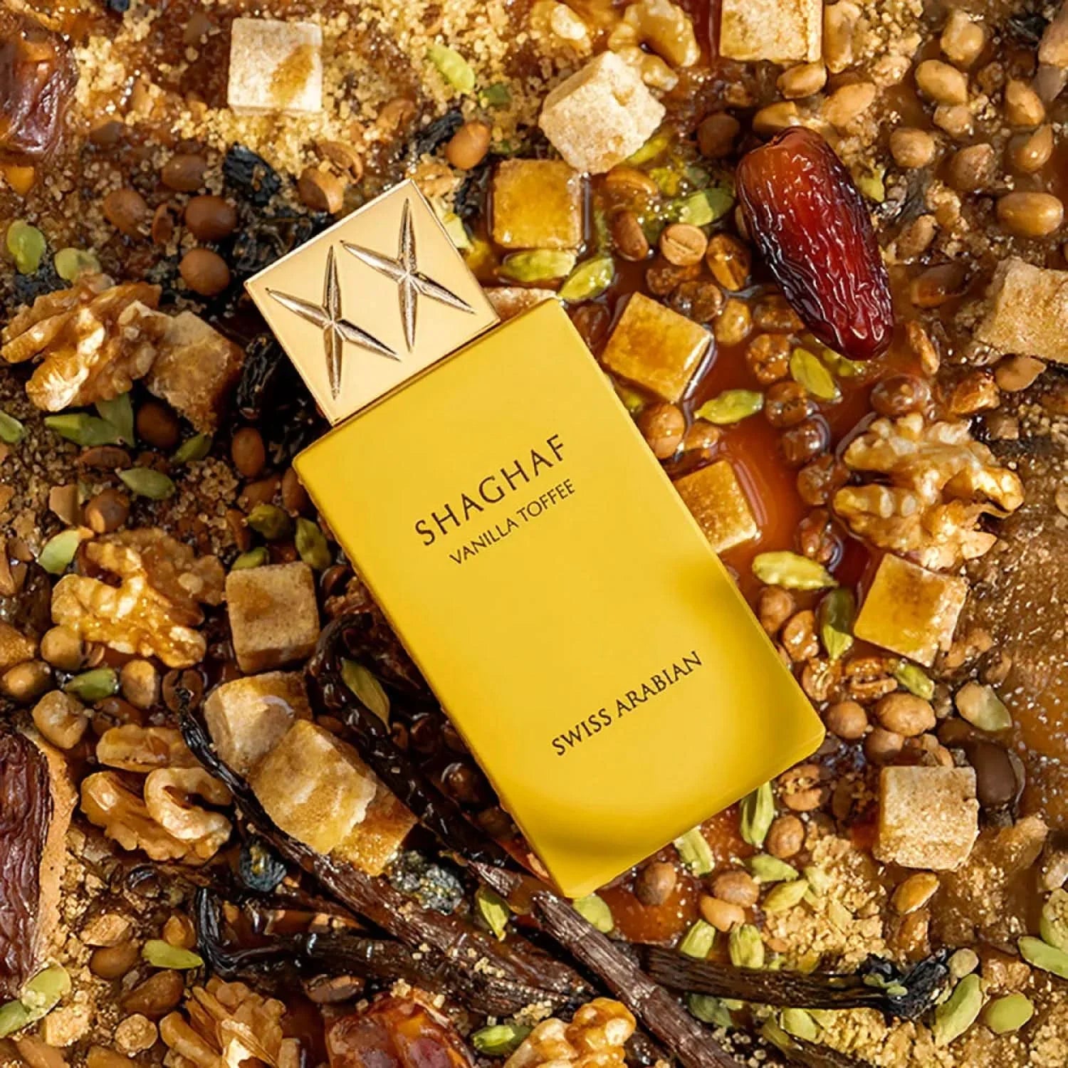 A yellow bottle of Swiss Arabian Shaghaf Vanilla Toffee EDP rests on a surface with nuts, dates, seeds, caramel pieces, and vanilla beans.