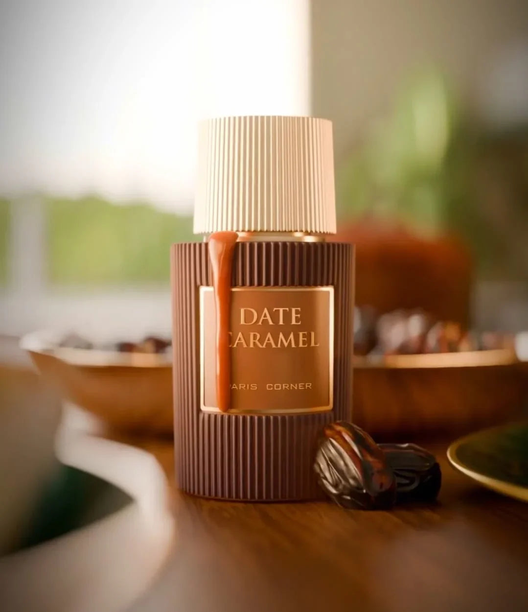 A bottle of Paris Corner Date Caramel 100ml Eau De Parfum sits on a table with caramel sauce dripping down, dates around it, and a soft green blurred background.
