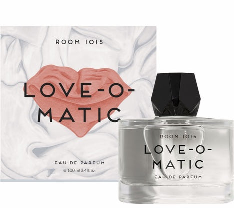 A round glass bottle of Room 1015 Love-O-Matic 100ml EDP sits in front of its matching white box, which features a pink heart graphic and the text “Love-O-Matic.”