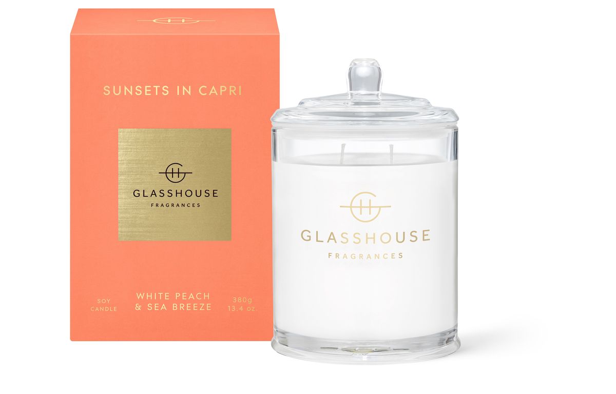 The Glasshouse Sunsets in Capri Soy Candle 380g features a "White Peach & Sea Breeze" label and comes in a transparent jar with a lid. It is accompanied by an orange box displaying the name and a gold square logo.